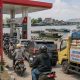 Ilustrasi SPBU di Palangka Raya dengan papan harga BBM, antrean kendaraan, truk distribusi sembako, serta sungai dan klotok di latar belakang.