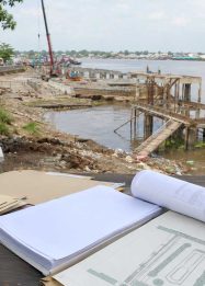 Ilustrasi proyek Waterfront City Kapuas dengan area tepi sungai, dermaga belum selesai, dan dokumen perencanaan pembangunan.